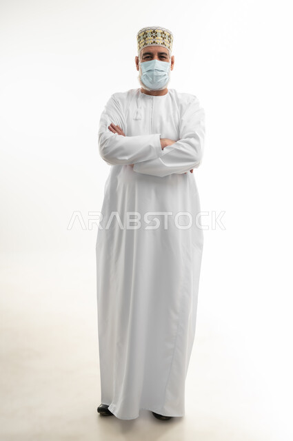 Standing with arms crossed in self-confidence gesture, taking precautions to prevent the spread of infection, portrait of an elderly Arab Gulf Omani man wearing a dishdasha and a kummah and wearing a medical mask, taking necessary measures to prevent the Corona virus, full body, white background