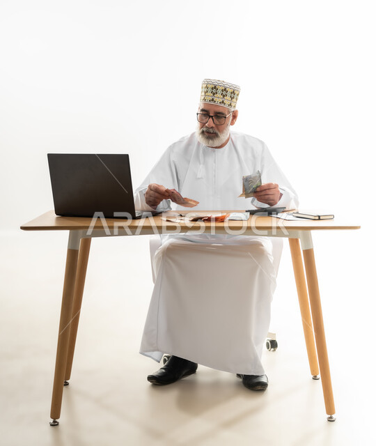الدفع والشراء أونلاين بالاعتماد على التقنيات الحديثة، جني الأموال باستخدام الحاسوب المحمول عبر العمل عن بعد، بورتريه لرجل شايب عربي خليجي عماني يرتدي الدشداشة والكمه يجلس خلف طاولة المكتب يقوم بعد النقود ويجري الحسابات، الجسم بالطول الكامل، خلفية بيضاء