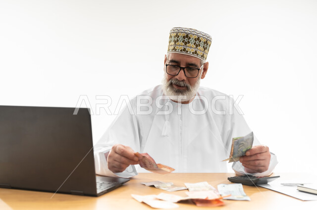 عد النقود وإجراء الحسابات المالية المهمة، الأعمال ذات الربح العالي عبر الانترنت، استخدام التقنيات والتكنولوجيا الحديثة للعمل أونلاين، بورتريه مقرب لرجل شايب عربي خليجي عماني يرتدي الدشداشة والكمه يجلس خلف طاولة المكتب وأمامه الحاسوب المحمول، خلفية بيضاء
