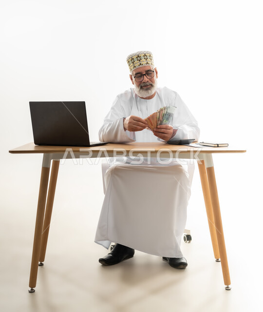 Making money using laptop by working remotely, relying on modern technologies in online payment and purchase, portrait of an old Arab Gulf Omani man wearing a dishdasha and a kummah sitting behind an office desk counting money and doing calculations, full body, white background