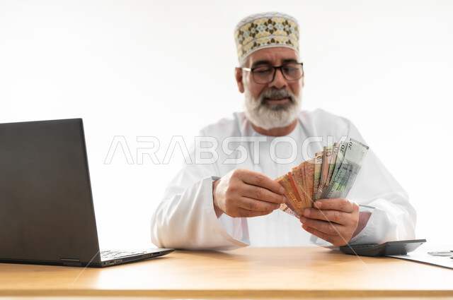 الأعمال ذات الربح العالي عبر الانترنت، عد النقود وإجراء الحسابات المالية المهمة، استخدام التقنيات والتكنولوجيا الحديثة للعمل أونلاين، بورتريه مقرب لرجل شايب عربي خليجي عماني يرتدي الدشداشة والكمه يجلس خلف طاولة المكتب وأمامه الحاسوب المحمول، خلفية بيضاء
