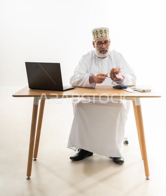 استخدام الحاسوب المحمول في جني الأموال عبر العمل عن بعد، الاعتماد على التقنيات الحديثة في الدفع والشراء أونلاين، بورتريه لرجل شايب عربي خليجي عماني يرتدي الدشداشة والكمه يجلس خلف طاولة المكتب يقوم بعد النقود ويجري الحسابات، الجسم بالطول الكامل، خلفية بيضاء