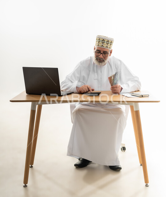 الاعتماد على التقنيات الحديثة في الدفع والشراء أونلاين، استخدام الحاسوب المحمول في جني الأموال عبر العمل عن بعد، بورتريه لرجل شايب عربي خليجي عماني يرتدي الدشداشة والكمه يجلس خلف طاولة المكتب يقوم بعد النقود ويجري الحسابات، الجسم بالطول الكامل، خلفية بيضاء