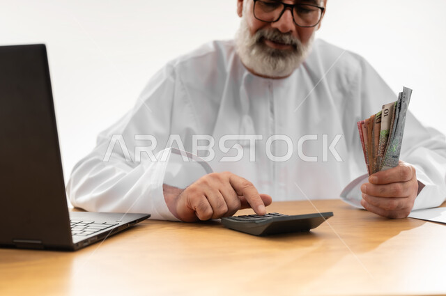 إجراء الحسابات المالية المهمة وعد النقود، الأعمال ذات الربح العالي عبر الانترنت، استخدام التقنيات والتكنولوجيا الحديثة للعمل أونلاين، بورتريه مقرب لرجل شايب عربي خليجي عماني يرتدي الدشداشة والكمه يجلس خلف طاولة المكتب وأمامه الحاسوب المحمول، خلفية بيضاء