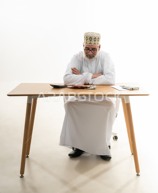 إيماءات الاحباط في محاولة الموازنة بين الراتب والمصروف، القيام بالحسابات الشهرية باستخدام الآلة الحاسبة، بورتريه لرجل شايب عربي خليجي عماني يرتدي الدشداشة والكمه والنظارة يجلس خلف المكتب متكتف اليدين وينظر للأوراق أمامه، الجسم بالطول الكامل، خلفية بيضاء