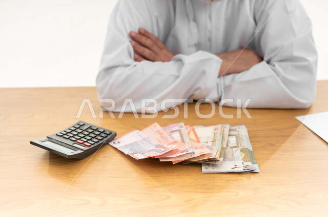 الجلوس بيدين مكتفتين بإيماءات الاحباط، محاولة الموازنة بين الراتب والمصروف، القيام بالحسابات الشهرية باستخدام الآلة الحاسبة، بورتريه مقرب لرجل شايب عربي خليجي عماني يرتدي الدشداشة يجلس خلف طاولة المكتب أمامه عدة فئات من الأوراق النقدية العمانية، خلفية بيضاء