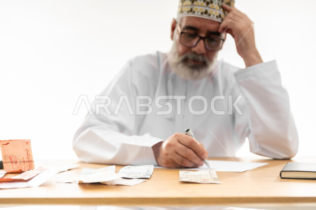 وضع اليد على الرأس بإيماءات التفكير والاحباط، القيام بالحسابات الشهرية باستخدام الآلة الحاسبة، محاولة الموازنة بين الراتب والمصروف، بورتريه مقرب  لرجل شايب عربي خليجي عماني يرتدي الدشداشة والكمه والنظارة يجلس خلف المكتب ويقوم بجمع وتوزيع النقود، خلفية بيضاء