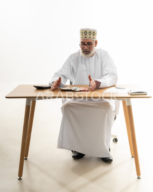 Doing monthly calculations using digital calculator, collecting money and trying to save and save, portrait of an old Arab Gulf Omani man wearing a dishdasha, a sleeve and glasses sitting behind an office table and collecting banknotes in his hands, full body, white background