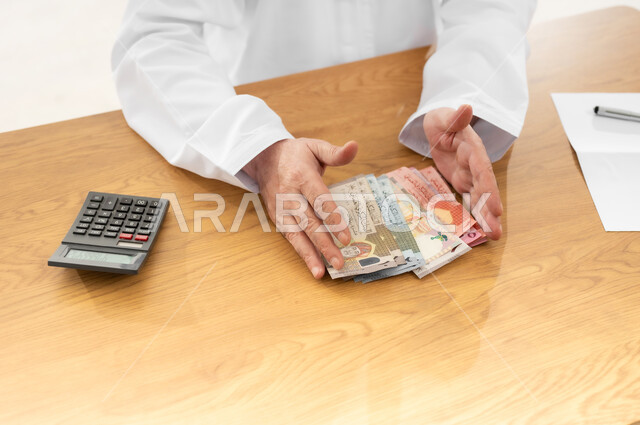 جمع النقود والقيام بالحسابات الشهرية باستخدام الآلة الحاسبة الرقمية، محاولة الادخار والتوفير، بورتريه مقرب لرجل شايب عربي خليجي عماني يرتدي الدشداشة التقليدية يجلس خلف طاولة المكتب ويجمع أوراق نقدية من عدة فئات بين يديه، خلفية بيضاء