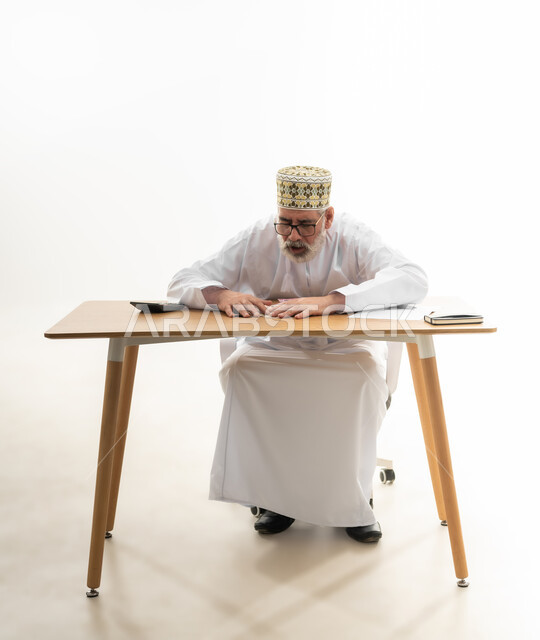 جمع النقود والقيام بالحسابات الشهرية باستخدام الآلة الحاسبة الرقمية، محاولة الادخار والتوفير، بورتريه لرجل شايب عربي خليجي عماني يرتدي الدشداشة والكمه والنظارة الطبية يجلس خلف طاولة المكتب ويجمع الأوراق النقدية بين يديه، الجسم بالطول الكامل، خلفية بيضاء