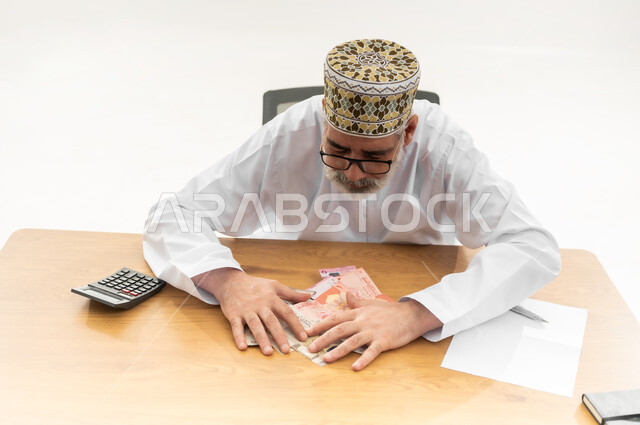 محاولة الادخار والتوفير، جمع النقود والقيام بالحسابات الشهرية باستخدام الآلة الحاسبة الرقمية، بورتريه مقرب لرجل شايب عربي خليجي عماني يرتدي الدشداشة التقليدية والكمه والنظارة الطبية يجلس خلف طاولة المكتب ويجمع أوراق نقدية من عدة فئات بين يديه، خلفية بيضاء