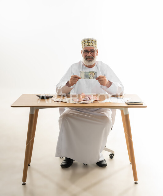 العملة الرسمية المتداولة في أسواق سلطنة عمان، عرض الإصدار الجديد للنقود الورقية العمانية، بورتريه لرجل شايب عربي خليجي عماني يرتدي الدشداشة والكمه والنظارة الطبية يجلس خلف طاولة المكتب ويمسك ورقة نقدية من فئة العشرين ريال، الجسم بالطول الكامل، خلفية بيضاء