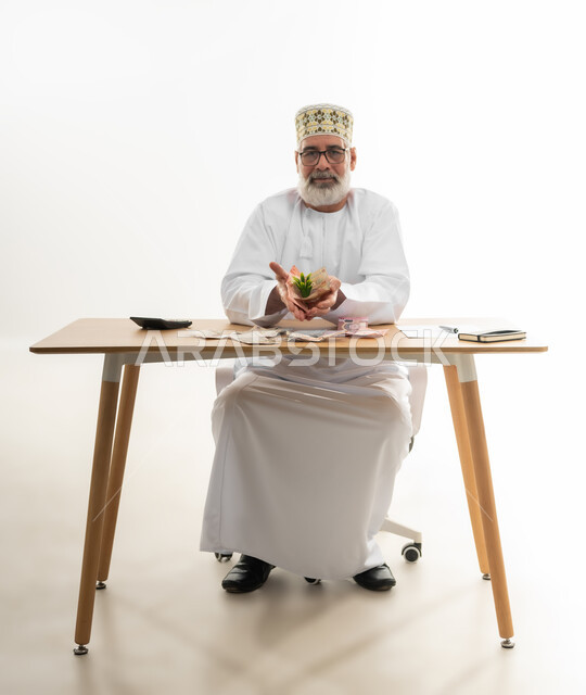 النمو والنهضة بالاقتصاد العماني، النظر إلى الكاميرا بإيماءات السعادة والسرور، بورتريه لرجل كبير بالسن عربي خليجي عماني يرتدي الدشداشة والكمه العمانية والنظارة الطبية يمسك نبتة خضراء تتوسط النقود الورقية في يديه، الجسم بالطول الكامل، خلفية بيضاء