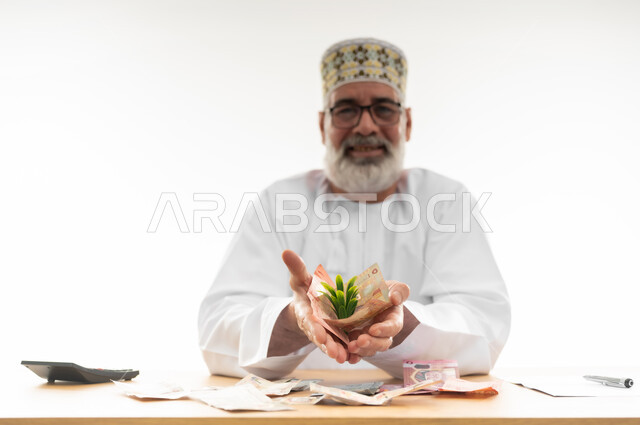 النظر إلى الكاميرا بإيماءات السعادة والسرور، النمو والنهضة بالاقتصاد العماني، بورتريه مقرب لرجل كبير بالسن عربي خليجي عماني يرتدي الدشداشة والكمه العمانية والنظارة الطبية يمسك نبتة خضراء تتوسط النقود الورقية في يديه، خلفية بيضاء