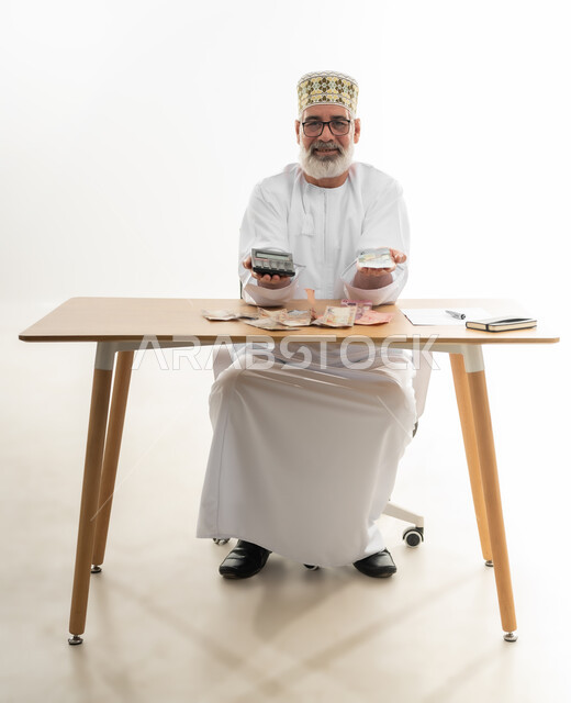 السعي نحو التوفير والادخار، محاولة إدارة المصروف المنزلي بتدبر، استخدام الحاسبة الرقمية في الأعمال الاقتصادية، بورتريه مقرب لرجل كبير بالسن عربي خليجي عماني يرتدي الدشداشة والكمه يجلس ويقوم بالحسابات الشهرية المالية، الجسم بالطول الكامل، خلفية بيضاء