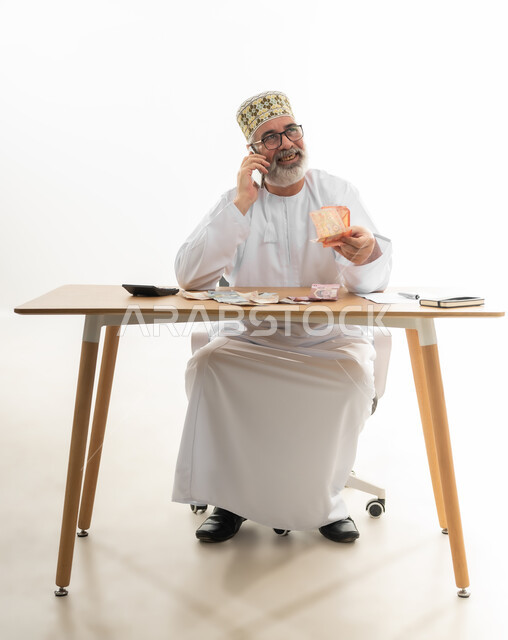 السؤال عن سعر شيء ما بإيماءات الأمل، الاستعلام عن الفواتير عبر جهاز الجوال، بورتريه لرجل كبير بالسن عربي خليجي عماني يرتدي الدشداشة والكمه والنظارة الطبية ويمسك النقود الورقية بيده، إجراء مكالمة هاتفية باستخدام الهاتف المحمول، الجسم بالطول الكامل، خلفية بيضاء