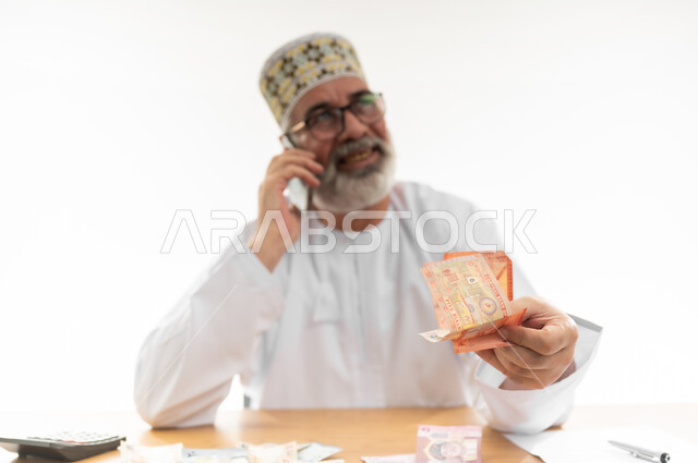 الاستعلام عن الفواتير عبر جهاز الجوال، السؤال عن سعر شيء ما بإيماءات الأمل، بورتريه مقرب لرجل كبير بالسن عربي خليجي عماني يرتدي الدشداشة والكمه العمانية يضع النظارة الطبية و يمسك النقود الورقية بيده، إجراء مكالمة هاتفية باستخدام الهاتف المحمول، خلفية بيضاء