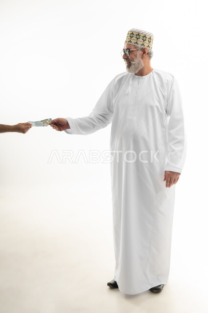 محاولة الاقتصاد في المصروف المنزلي، ادخار النقود لوقت الحاجة، بورتريه لرجل كبير بالسن عربي خليجي عماني يرتدي الدشداشة والكمه العمانية التقليدية ويضع النظارة الطبية يقوم بدفع الفواتير وحسابات المشتريات، الجسم بالطول الكامل، خلفية بيضاء