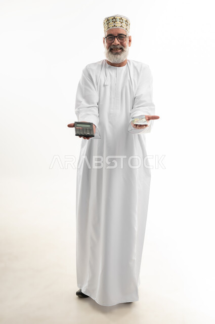 محاولة إدارة المصروف المنزلي بتدبر، استخدام الحاسبة الرقمية في الأعمال الاقتصادية، بورتريه لرجل كبير بالسن عربي خليجي عماني يرتدي الدشداشة والكمه العمانية التقليدية يقوم بالحسابات بإيماءات التركيز، السعي نحو التوفير والادخار، الجسم بالطول الكامل، خلفية بيضاء