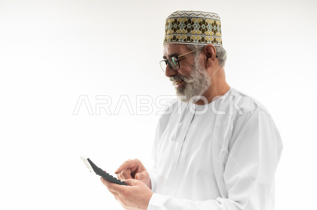 استخدام الحاسبة الرقمية في الأعمال الاقتصادية المالية، محاولة إدارة المصروف المنزلي بتدبر، بورتريه مقرب لرجل كبير بالسن عربي خليجي عماني يرتدي الدشداشة والكمه العمانية التقليدية يقوم بالحسابات بإيماءات التركيز، السعي نحو التوفير والادخار، خلفية بيضاء