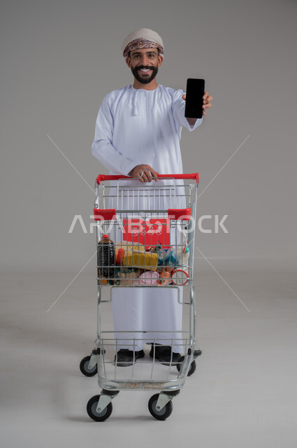 الاستمتاع بالتسوق في المولات والمراكز التجارية، شراء المستلزمات والحاجيات الضرورية، الاستفادة من الخصومات على المنتجات، بورتريه لرجل عربي خليجي عماني يرتدي الدشداشة والكمه ويجر عربة التسوق أمامه ويقوم بعرض شاشة سوداء فارغة، الجسم بالطول الكامل، خلفية بيضاء 