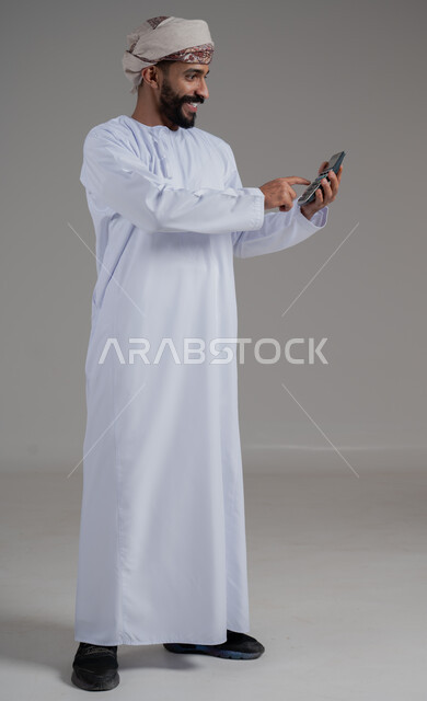 مفهوم إدارة الشؤون الاقتصادية، العمل في إدارة الحسابات، بورتريه لرجل عربي خليجي عماني يرتدي الدشداشة والعمامة يشير بإصبع السبابة إلى الآلة الحاسبة الرقمية، توفير ودراسة الأمور المالية, الوقوف باستقامة بايماءات السعادة والبشاشة، الطول الكامل للجسم، خلفية رمادية