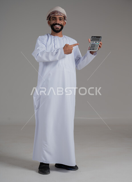 العمل في إدارة الحسابات، بورتريه لرجل عربي خليجي عماني يرتدي الدشداشة والعمامة يشير بإصبع السبابة إلى الآلة الحاسبة الرقمية، توفير ودراسة الأمور المالية, النظر الى الكاميرا بايماءات السعادة والبشاشة، مفهوم إدارة الشؤون الاقتصادية، الطول الكامل للجسم، خلفية رمادية