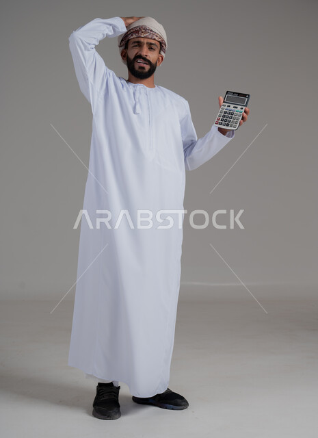 الإحصائيات والقروض البنكية, استخدام الآلة الحاسبة في إجراء العمليات الحسابية، العمل في إدارة الحسابات، بورتريه لرجل عربي خليجي عماني يرتدي الدشداشة والعمامة يضع يده على رأسه بايماءات الانزعاج والاستغراب، الطول الكامل للجسم، خلفية رمادية