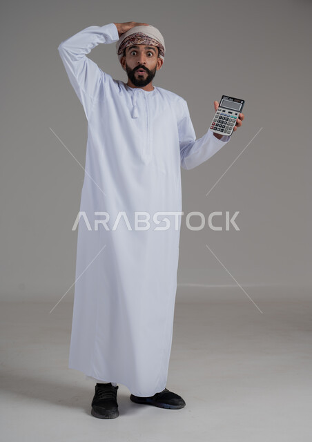 استخدام الآلة الحاسبة في إجراء العمليات الحسابية، العمل في إدارة الحسابات، بورتريه لرجل عربي خليجي عماني يرتدي الدشداشة والعمامة يضع يده على رأسه بايماءات الذهول الدهشة، الإحصائيات والقروض البنكية, الطول الكامل للجسم، خلفية رمادية