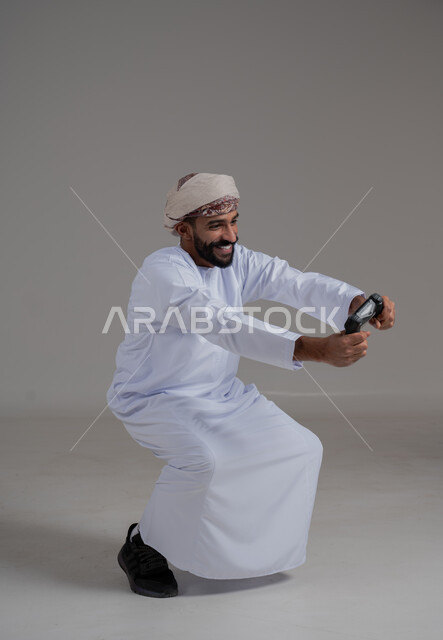 تجربة ترفيهية ممتعة ومثيرة في الأجهزة الحديثة، ايماءات السعادة والسرور، التفاعل والاندماج مع ألعاب البلايستيشن، تقنيات الكترونية ترفيهيه, بورتريه لرجل عربي ععماني خليجي يرتدي الدشداشة والعمامة يحمل بيده عصا التحكم، خلفية رمادية