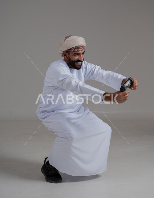 تقنيات الكترونية ترفيهيه, بورتريه لرجل عربي ععماني خليجي يرتدي الدشداشة والعمامة يحمل بيده عصا التحكم، تجربة ترفيهية ممتعة ومثيرة في الأجهزة الحديثة، ايماءات السعادة والسرور، التفاعل والاندماج مع ألعاب البلايستيشن، خلفية رمادية