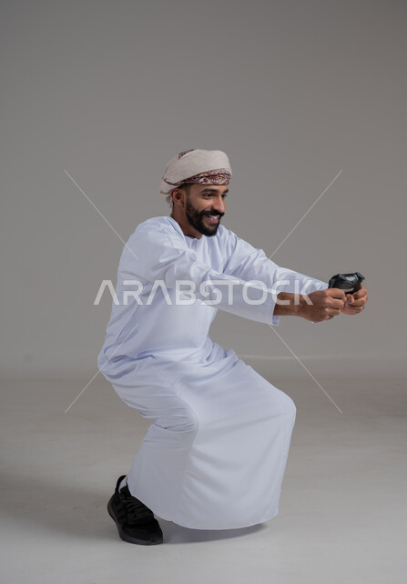 استخدام الأجهزة التكنولوجية والتقنيات الحديثة، بورتريه لرجل عربي ععماني خليجي يرتدي الدشداشة والعمامة يحمل بيده عصا التحكم بإيماءات التفاعل والاندماج، تجربة ترفيهية ممتعة ومثيرة، قضاء وقت ممتع بالألعاب الالكترونية، الطول الكامل للجسم، خلفية رمادية  