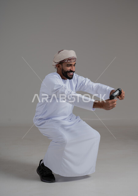 Having fun with electronic games, using technological devices and modern techniques, portrait of an Arab Omani Gulf man wearing a dishdasha and turban holding a joystick with gestures of interaction and integration, a fun and exciting entertainment experience, full body length, gray background