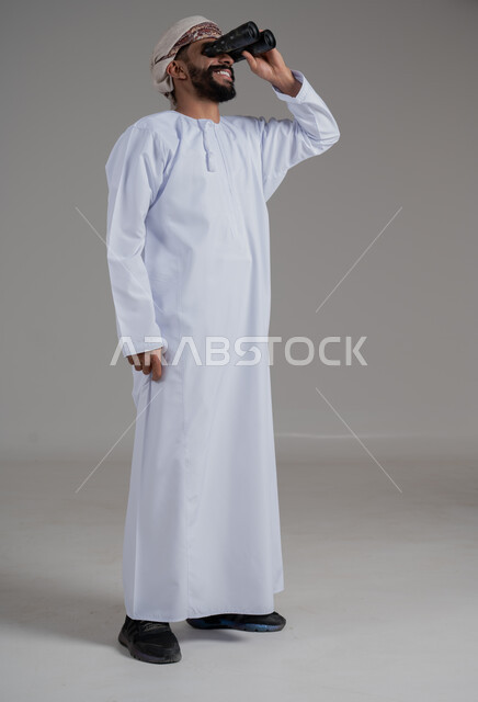 Seeing distant objects and bringing them closer, portrait of an Arab Gulf Omani man wearing a dishdasha and turban holding binoculars in his hand looking at something with gestures of pleasure and amazement, concept of future outlook, love of exploration and adventure, full body length, gray background
