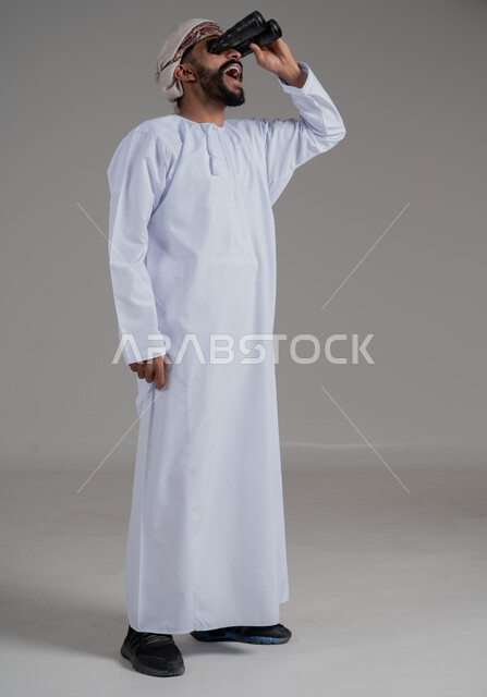 حب الاستكشاف و المغامرة, رؤية الاشياء البعيدة و تقريبها, بورتريه لرجل عربي خليجي عماني يرتدي الدشداشة والعمامة يحمل بيده المنظار ثنائي العينين ينظر الى شيء ما بايماءات السرور والذهول، مفهوم النظرة المستقبلية، الطول الكامل للجسم، خلفية رمادية