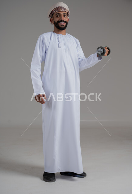 نشاط بدني للمحافظة على الصحة و اللياقة الجسدية، بناء عضلات الجسم وتقويتها، رفع الأوزان الحديدية الثقيلة، بورتريه لرجل عربي خليجي عماني يرتدي الدشداشة والعمامة يحمل بيده الدمبلز، النظرالى الكاميرا بايماءات السعادة والسرور، الطول الكامل للجسم، خلفية رمادية
