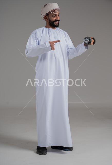 رفع الأوزان الحديدية الثقيلة، بورتريه لرجل عربي خليجي عماني يرتدي الدشداشة والعمامة يحمل بيده الأثقال، الاشارة باصبع السبابة الى الدامبلز، نشاط بدني للمحافظة على الصحة و اللياقة الجسدية، بناء عضلات الجسم وتقويتها، الطول الكامل للجسم، خلفية رمادية