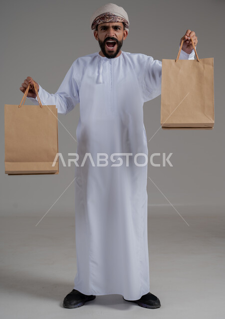 الإمساك بحقائب التسوق الورقية، النظر للكاميرا بإيماءات الاندهاش, مواسم التخفيضات والحسومات, بورتريه لشاب عربي خليجي عماني يرتدي الدشداشة والعمامة يعرض أكياس المشتريات التجارية والهدايا، التبضع وأخذ الحاجات الضرورية, جسم بالطول الكامل, خلفية رمادية