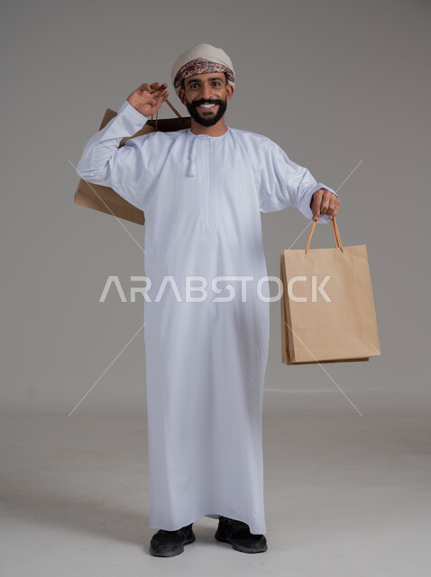 النظر الى الكاميرا بثقة و سعادة للاستفادة من العروض و التخفيضات, بورتريه لشاب عربي خليجي عماني مبتسم يرتدي الدشداشة والعمامة يحمل بيده اكياس التسوق الورقية, متعة في التسويق و اقتناء المستلزمات و الهدايا, صورة الجسم بالطول الكامل, خلفيه رمادية