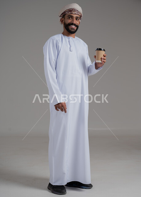 النظر للكاميرا والاستمتاع بتناول المشروبات المفضلة, احتساء مشروب ساخن لذيذ, بورتريه لشاب عربي خليجي عماني يرتدي الدشداشة والعمامة ويحمل بيده كوب من القهوة, البن العربي الفاخر الأصيل, التلذذ بمشاريب الكافيهات والمطاعم الجاهزة، الجسم بالطول الكامل، خلفية رمادية