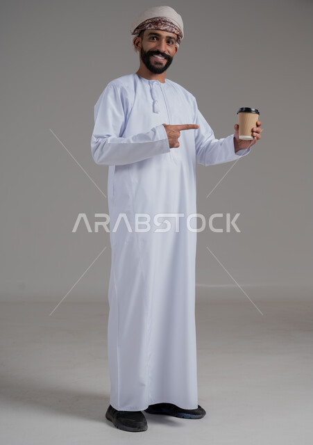 الاستمتاع بتناول المشروبات الساخنة والأعشاب الشتوية الدافئة، بورتريه لرجل عربي خليجي عماني يرتدي الدشداشة والعمامة يشير باصبع السبابة الى كوب ورقي مقوى من القهوة، تأثير الكافيين على نشاط الدماغ والذاكرة، صورة الجسم بالطول الكامل، خلفية رمادية