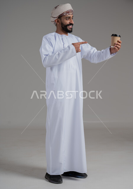 تأثير الكافيين على نشاط الدماغ والذاكرة، الاستمتاع بتناول المشروبات الساخنة والأعشاب الشتوية الدافئة، بورتريه لرجل عربي خليجي عماني يرتدي الدشداشة والعمامة يشير باصبع السبابة الى كوب ورقي مقوى من القهوة، صورة الجسم بالطول الكامل، خلفية رمادية