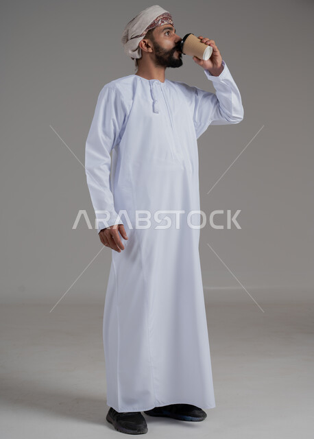 احتساء مشروب ساخن لذيذ, بورتريه لشاب عربي خليجي عماني يرتدي الدشداشة والعمامة ويحمل بيده كوب من القهوة, البن العربي الفاخر الأصيل, التلذذ بمشاريب الكافيهات والمطاعم الجاهزة، الاستمتاع بتناول المشروبات المفضلة, الجسم بالطول الكامل، خلفية رمادية