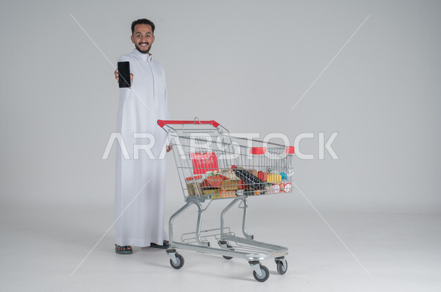 الاستمتاع بالتسوق في المولات والمراكز التجارية، شراء المستلزمات والحاجيات الضرورية، الاستفادة من الخصومات على المنتجات، بورتريه لرجل عربي خليجي سعودي يرتدي الثوب الابيض ويجر عربة التسوق أمامه ويقوم بعرض شاشة سوداء فارغة بحماس، الجسم بالطول الكامل، خلفية بيضاء 