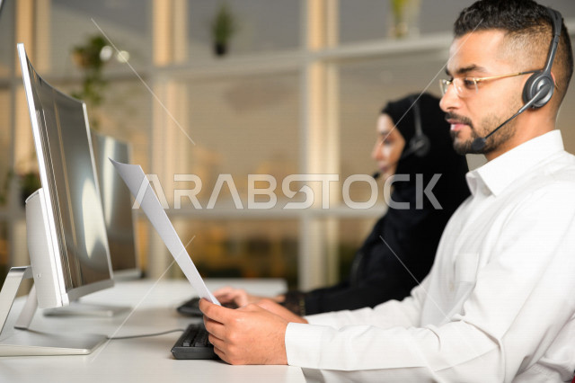 موظف عربي خليجي سعودي يعمل في خدمة العملاء ويمسك الاوراق بيده، موظف وموظفة يرتديان سماعة الاتصال، استعلامات واستفسارات، الاجابه على اسئلة العملاء، اجراء اتصال فيديو من خلال الحاسوب، متابعة وتطوير العمل