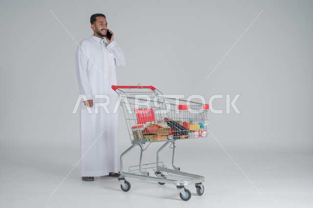 الاستمتاع بالتسوق في المولات والمراكز التجارية، شراء المستلزمات والحاجيات الضرورية، الاستفادة من الخصومات على المنتجات، بورتريه لرجل عربي خليجي سعودي يرتدي الثوب الابيض ويجر عربة التسوق أمامه ويقوم بإجراء مكالمة هاتفية، الجسم بالطول الكامل، خلفية بيضاء 
