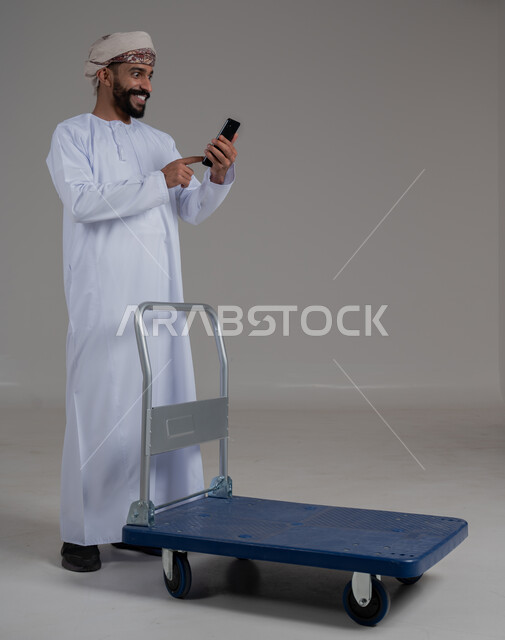 استخدام الموبايل لإنجاز المهام, ايماءات السعادة و السرور، عربة دفع يدوية لنقل الأغراض والأمتعة، بورتريه لرجل عربي خليجي عماني يرتدي الدشداشة والعمامة يحمل بيده الهاتف المحمول، التواصل مع الاهل و الاصدقاء, الطول الكامل للجسم،  خلفية رمادية