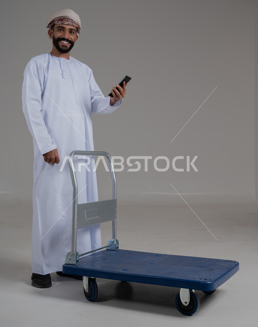 النظر الى الكاميرا بايماءات السعادة و السرور، عربة دفع يدوية لنقل الأغراض والأمتعة، بورتريه لرجل عربي خليجي عماني يرتدي الدشداشة والعمامة يحمل بيده الهاتف المحمول، إستخدام الموبايل لإنجاز المهام, التواصل مع الاهل و الاصدقاء, الطول الكامل للجسم،  خلفية رمادية