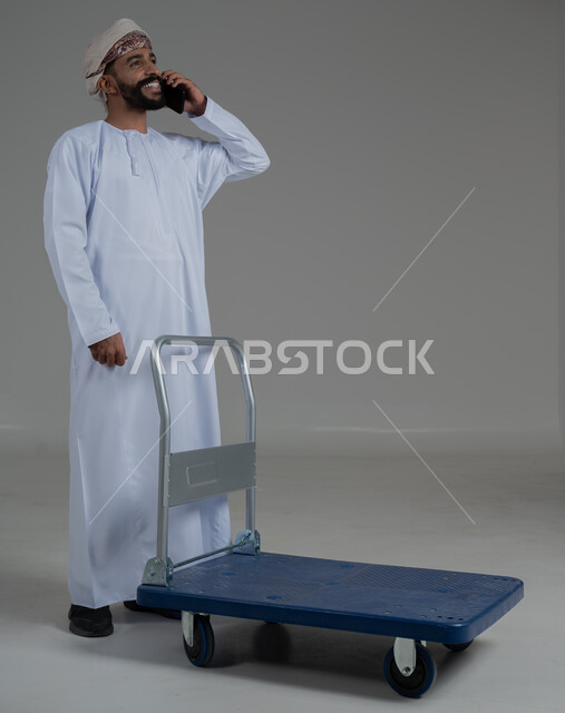 التواصل مع الاهل و الاصدقاء, ايماءات السعادة و السرور، عربة دفع يدوية لنقل الأغراض والأمتعة، بورتريه لرجل عربي خليجي عماني يرتدي الدشداشة والعمامة يقوم بإجراء مكالمة هاتفية عبر الهاتف المحمول، الطول الكامل للجسم،  خلفية رمادية