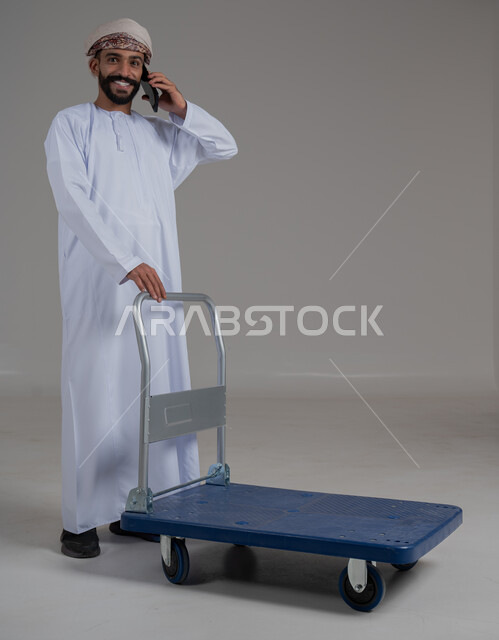 عربة دفع يدوية لنقل الأغراض والأمتعة، بورتريه لرجل عربي خليجي عماني يرتدي الدشداشة والعمامة يقوم بإجراء مكالمة هاتفية عبر الهاتف المحمول، التواصل مع الاهل و الاصدقاء, ايماءات السعادة و السرور، الطول الكامل للجسم،  خلفية رمادية